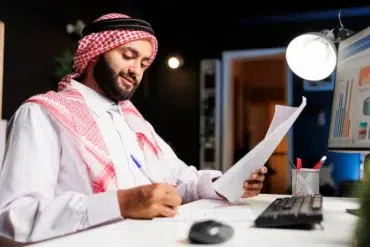 Man in traditional attire works at a desk, reviewing documents and jotting notes. A computer displaying charts is on, indicating a focused office setting.