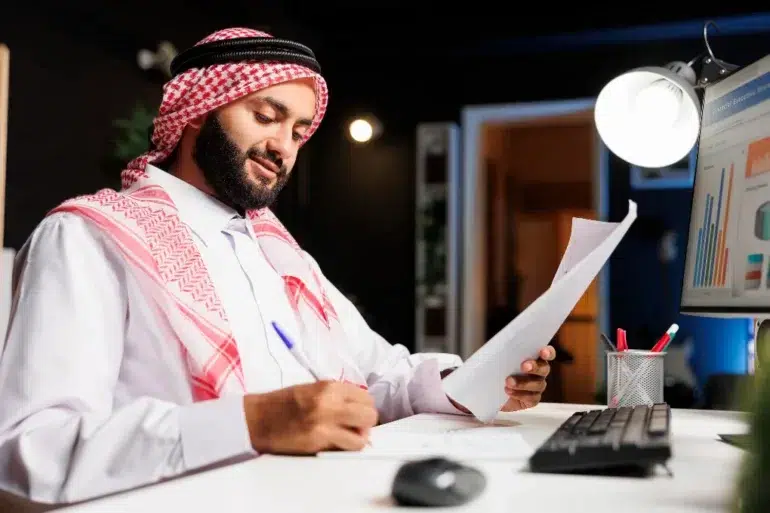 Man in traditional attire works at a desk, reviewing documents and jotting notes. A computer displaying charts is on, indicating a focused office setting.