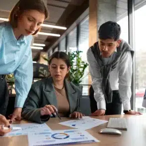 Three colleagues collaborate on documents in a modern office. A computer displays charts. The atmosphere is focused and professional.
