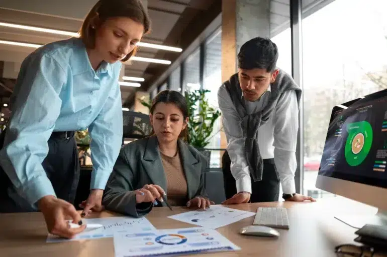Three colleagues collaborate on documents in a modern office. A computer displays charts. The atmosphere is focused and professional.