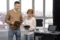 A man and woman stand in an office with large windows. The man holds a tablet and wears a brown sweater; the woman, holding a pen on a notepad, wears a white blouse. They appear focused and engaged in discussion. In the background, there are computers and cityscape views.