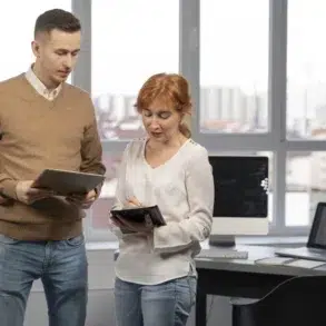 A man and woman stand in an office with large windows. The man holds a tablet and wears a brown sweater; the woman, holding a pen on a notepad, wears a white blouse. They appear focused and engaged in discussion. In the background, there are computers and cityscape views.