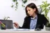 Person in business attire pointing at a laptop screen at a desk with documents, plants, and office supplies.
