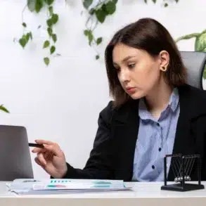 Person in business attire pointing at a laptop screen at a desk with documents, plants, and office supplies.