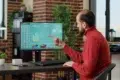 Man in red sweater sitting at desk, pointing at computer screen displaying charts and global stock statistics in a modern office.