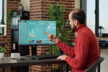 Man in red sweater sitting at desk, pointing at computer screen displaying charts and global stock statistics in a modern office.