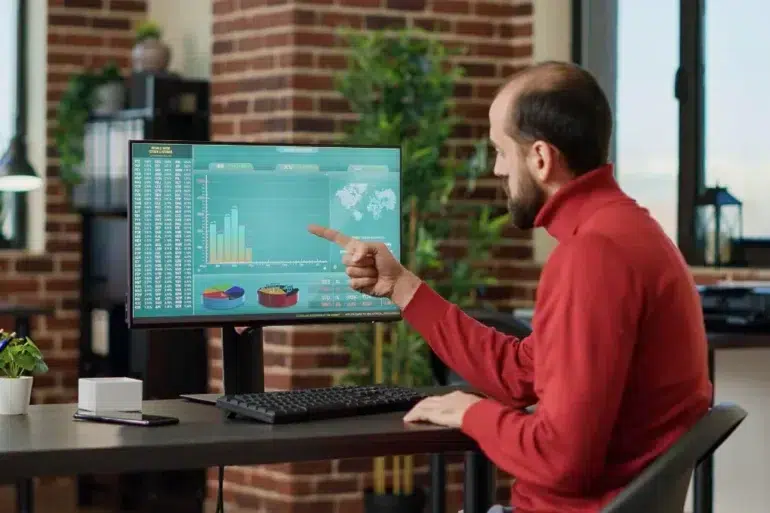 Man in red sweater sitting at desk, pointing at computer screen displaying charts and global stock statistics in a modern office.