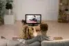 A couple sits on a couch, watching breaking news on a TV in a cozy living room. The tone is calm and focused, with a plant and shelves in view.