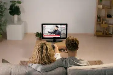 A couple sits on a couch, watching breaking news on a TV in a cozy living room. The tone is calm and focused, with a plant and shelves in view.