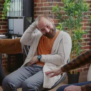 A group therapy session with three people: a woman taking notes, a man smiling and scratching his head, and an older man gesturing, set against a cozy, brick-walled room.