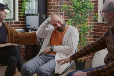 A group therapy session with three people: a woman taking notes, a man smiling and scratching his head, and an older man gesturing, set against a cozy, brick-walled room.