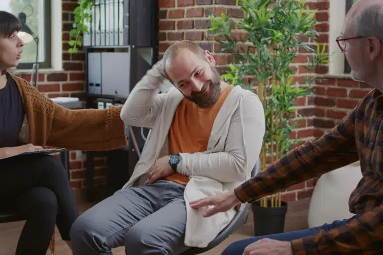 A group therapy session with three people: a woman taking notes, a man smiling and scratching his head, and an older man gesturing, set against a cozy, brick-walled room.