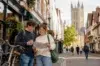 Two students stand on a charming historic street in Canterbury, smiling as they check a paper map, with bicycles, quaint shopfronts, and Canterbury Cathedral rising in the background.