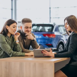 A couple sits with a saleswoman at a dealership, reviewing information on a tablet. They appear engaged and thoughtful, surrounded by cars.