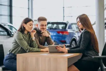 A couple sits with a saleswoman at a dealership, reviewing information on a tablet. They appear engaged and thoughtful, surrounded by cars.