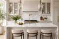 Spacious kitchen with marble countertops and island, featuring three cushioned stools, gold fixtures, pendant lights, and greenery, creating an elegant, airy feel.