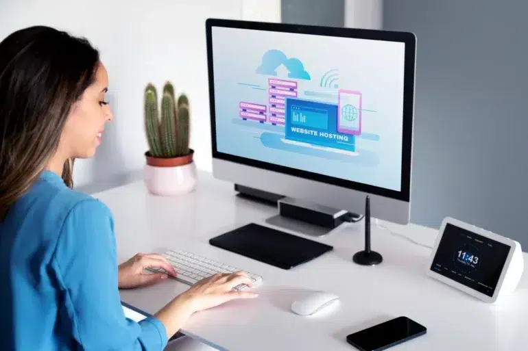 A woman in a blue shirt works at a computer with a website hosting graphic. The desk holds a tablet, smartphone, speaker, and a cactus. The atmosphere is modern and focused.
