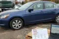 A blue sedan parked in a lot with laptops displaying a VIN and vehicle history report. Overcast sky and autumn trees set a somber, investigative tone.