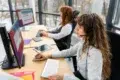 Two women working in a bright office, wearing headsets, focusing on computer screens. Their environment is professional and tech-driven.
