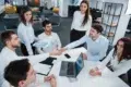 Office meeting scene with six diverse individuals around a table. Two men shake hands, symbolizing agreement. Open laptop and papers suggest collaboration.