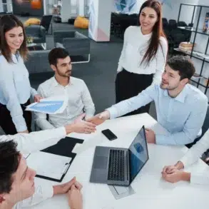 Office meeting scene with six diverse individuals around a table. Two men shake hands, symbolizing agreement. Open laptop and papers suggest collaboration.