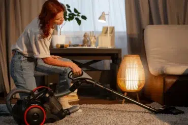 A woman vacuuming a cozy living room with a modern vacuum cleaner. Warm lighting from a lamp and a desk create an inviting atmosphere.