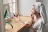 A woman in a robe and towel turban applies face cream at a wooden vanity. Skincare products are arranged beside her, with a potted plant in view.