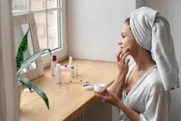 A woman in a robe and towel turban applies face cream at a wooden vanity. Skincare products are arranged beside her, with a potted plant in view.