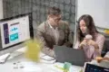 A man in a suit and a woman holding a coffee cup collaborate at a desk with a laptop. A computer screen displays graphs, indicating a professional setting.