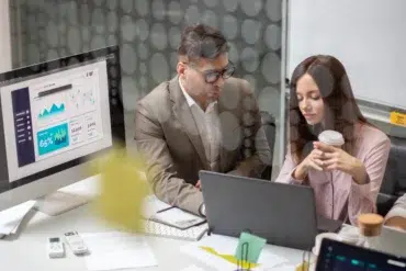 A man in a suit and a woman holding a coffee cup collaborate at a desk with a laptop. A computer screen displays graphs, indicating a professional setting.