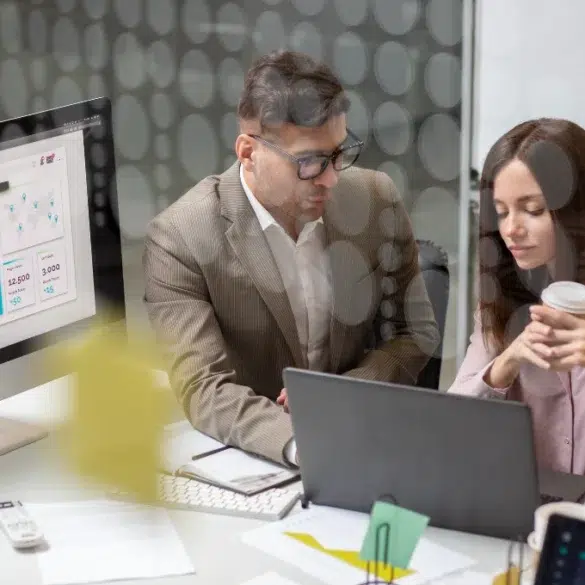 A man in a suit and a woman holding a coffee cup collaborate at a desk with a laptop. A computer screen displays graphs, indicating a professional setting.