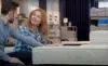 A couple sits in a store, inspecting a mattress. The woman, in a blue plaid shirt, smiles at the man, setting a warm, relaxed tone.