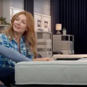 A couple sits in a store, inspecting a mattress. The woman, in a blue plaid shirt, smiles at the man, setting a warm, relaxed tone.