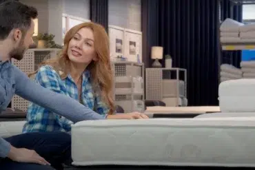 A couple sits in a store, inspecting a mattress. The woman, in a blue plaid shirt, smiles at the man, setting a warm, relaxed tone.