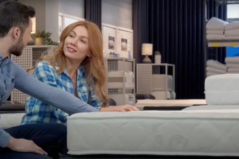 A couple sits in a store, inspecting a mattress. The woman, in a blue plaid shirt, smiles at the man, setting a warm, relaxed tone.