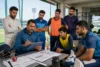 A cricket coach discusses strategy on a tablet with attentive players in a sports facility, creating a focused and analytical atmosphere.