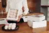 A person in a white sweater holds a box of pink meringues in a kitchen. Nearby, more meringue boxes and a plate with meringues evoke a cozy, homemade feel.