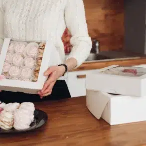 A person in a white sweater holds a box of pink meringues in a kitchen. Nearby, more meringue boxes and a plate with meringues evoke a cozy, homemade feel.