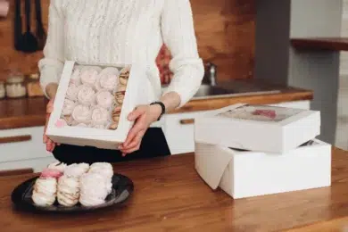 A person in a white sweater holds a box of pink meringues in a kitchen. Nearby, more meringue boxes and a plate with meringues evoke a cozy, homemade feel.