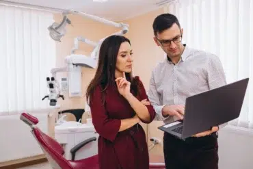 Two healthcare professionals reviewing information on a laptop in a dental clinic with equipment in the background.