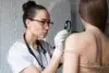 A focused female doctor in a white coat and gloves examines a mole on a patient's back using a magnifying glass, in a clinical setting.