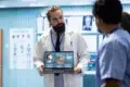 A doctor with a beard in a white coat holds a laptop displaying medical data, presenting to colleagues in a clinical setting. The mood is focused and professional.