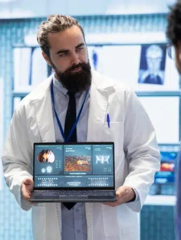 A doctor with a beard in a white coat holds a laptop displaying medical data, presenting to colleagues in a clinical setting. The mood is focused and professional.