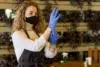 A woman wearing a black face mask and white blouse is putting on blue gloves. She wears a dark apron, standing in an indoor setting with potted plants.