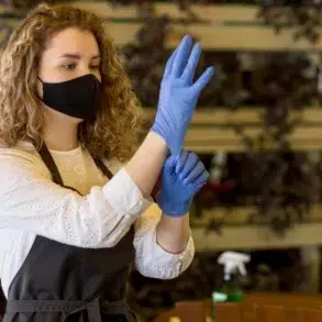 A woman wearing a black face mask and white blouse is putting on blue gloves. She wears a dark apron, standing in an indoor setting with potted plants.