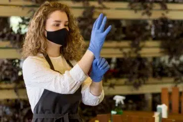 A woman wearing a black face mask and white blouse is putting on blue gloves. She wears a dark apron, standing in an indoor setting with potted plants.