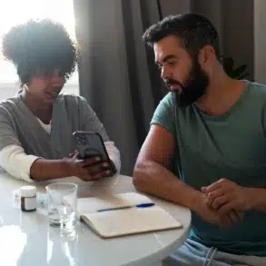 A nurse and a man sit at a table. The nurse shows him a smartphone screen, with a notebook, pill bottles, glass of water, and apples nearby.