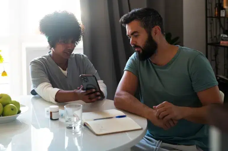 A nurse and a man sit at a table. The nurse shows him a smartphone screen, with a notebook, pill bottles, glass of water, and apples nearby.