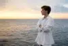 Person in a white shirt stands with arms crossed, looking out over a calm sea at sunset under a partly cloudy sky.
