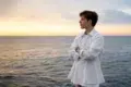 Person in a white shirt stands with arms crossed, looking out over a calm sea at sunset under a partly cloudy sky.